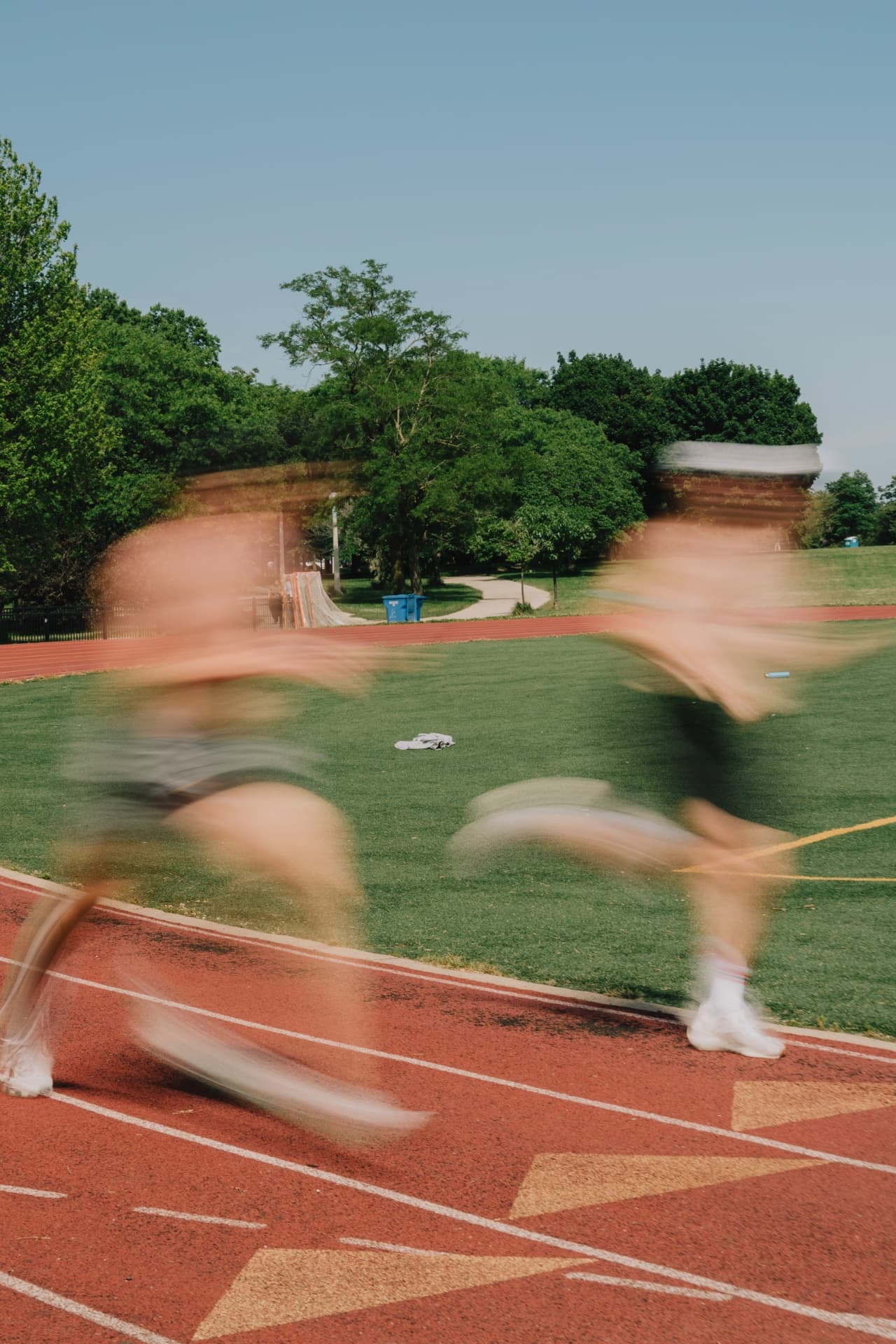 Two runners sprinting on a red track with heavy motion blur, green field and trees behind under a clear blue sky.