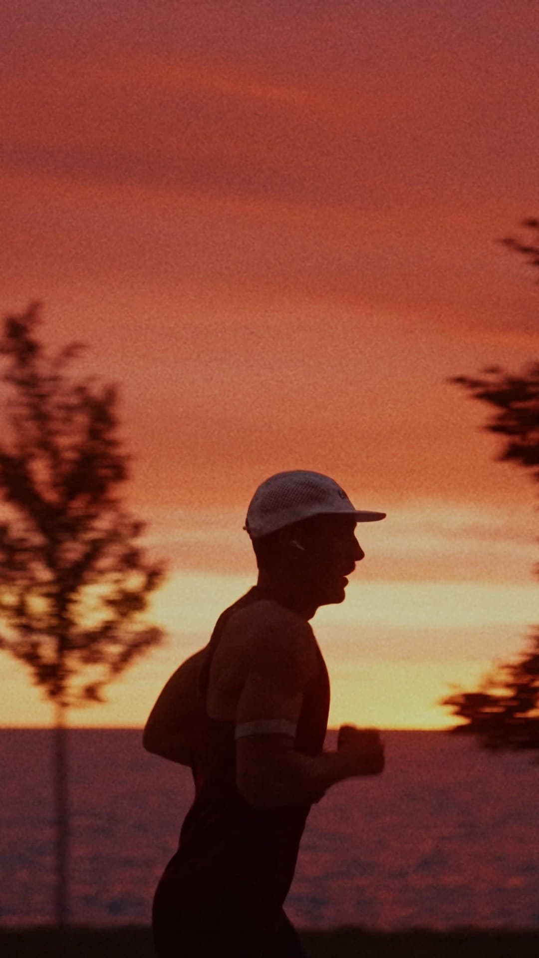 Silhouetted runner in profile wearing a cap, jogging along a shoreline with orange-red sunset sky and blurred trees behind.