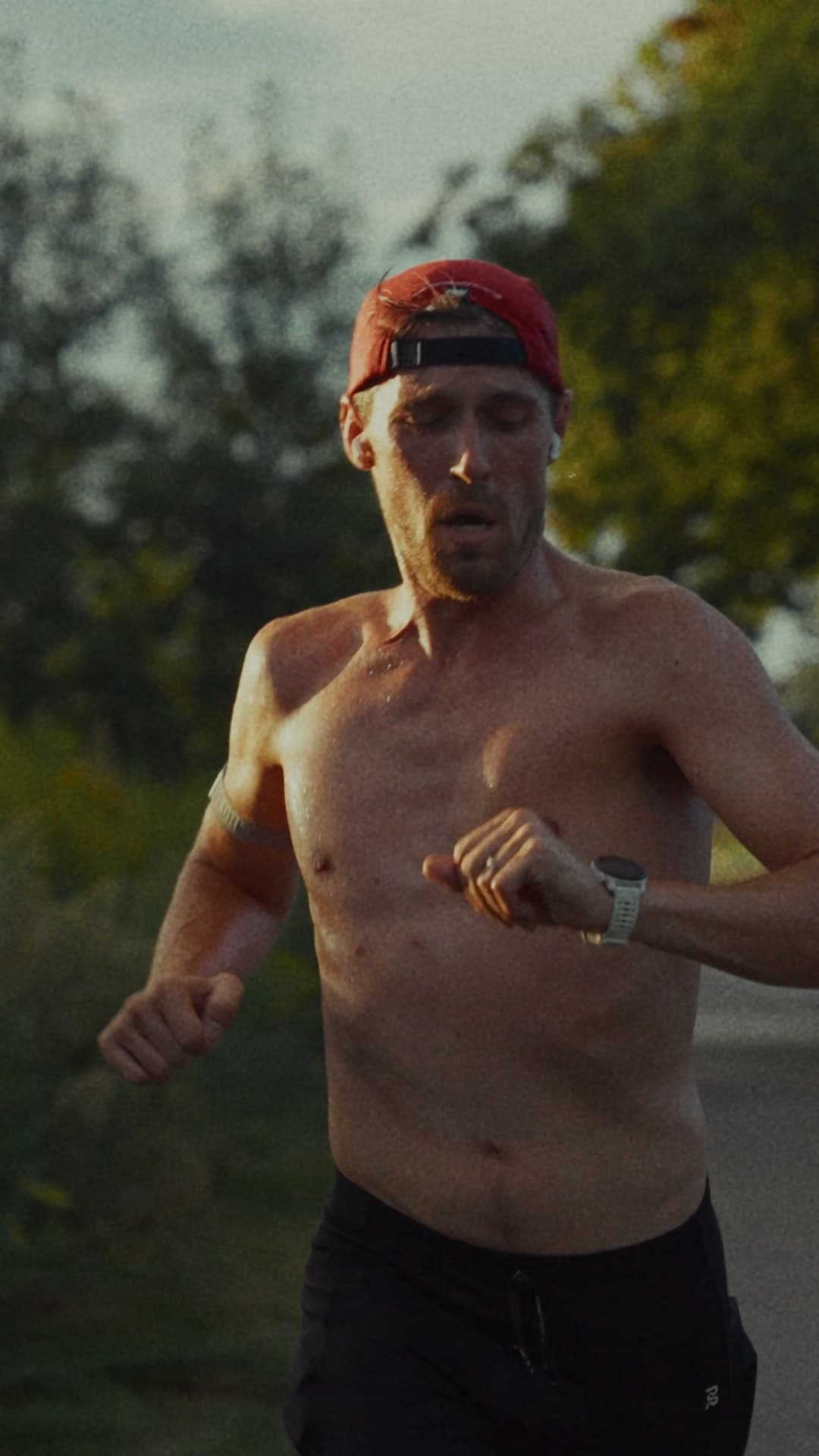 Shirtless male runner wearing a red backwards cap and white watch, running on a road with trees and warm evening light.