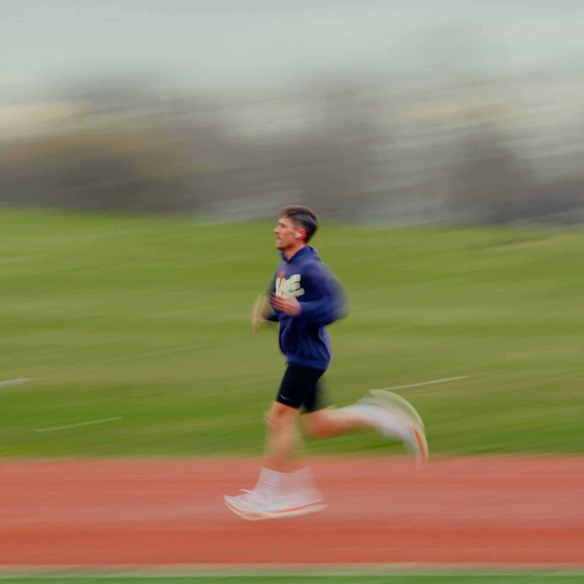 A male runner in a blue hoodie and black shorts sprinting along a red track, captured with side panning motion blur against a grassy background.